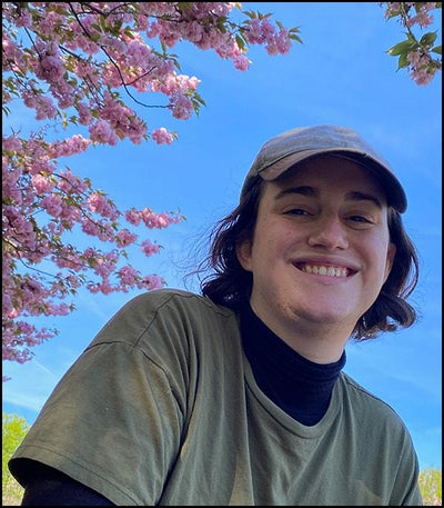 Sam Leander looking down and smiling at the camera wearing an olive green t-shirt, grey ball cap, and a beautiful bright blue sky and cherry blossom blooming int he background