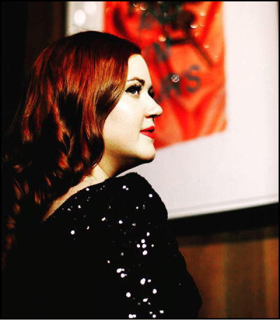 Sarah Jayne Kinney side profile shot wearing a sparkly black long sleeve, curled red hair, and red lipstick, with a red artwork blurred in the background