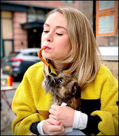 Polly Rodriguez headshot in a pink sweater with pasta in her mouth and puppy in her lap lady and the Tramp romance style with NYC street blurred in the background