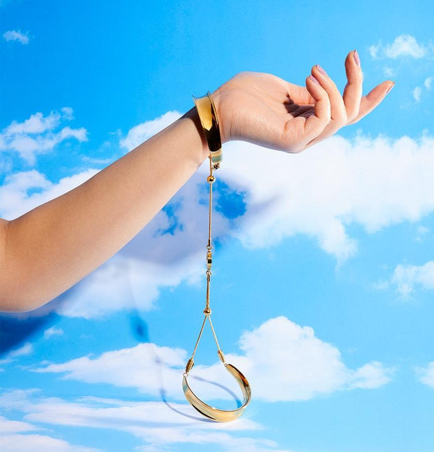 Gold bangle on a models wrist on a bright blue background with white clouds